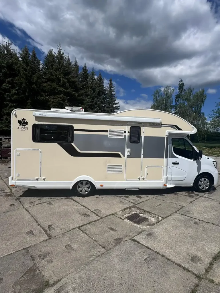 Beige-weißes Ahorn Canada Wohnmobil, seitlich fotografiert auf einem Parkplatz mit Bäumen im Hintergrund.