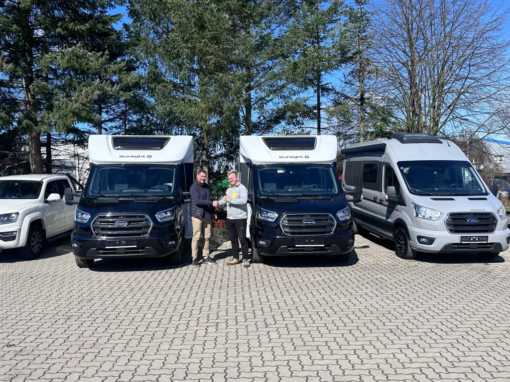 Drei Wohnmobile der Marke Bürstner nebeneinander auf einem Parkplatz, zwei schwarze und ein silbernes Modell, davor zwei Personen bei der Fahrzeugübergabe.